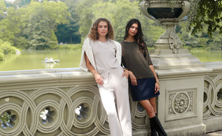 Two women standing on a stone bench in a park with trees and water in the background.